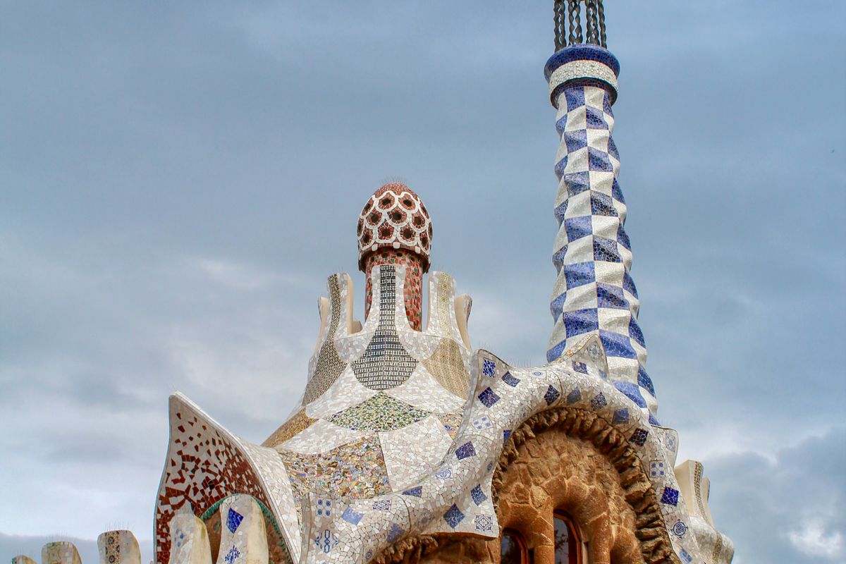 Close view of a Park Güell gatehouse roof in broken-tile trencadís mosaic, with a tall blue-and-white tiled spire rising against a grey sky